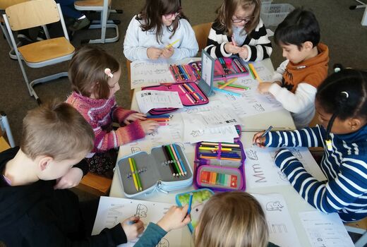 Mehrere Kinder sitzen gemeinsam an Tischen. Sie schreiben auf einem Arbeitsblatt mit Buntstiften den Groß- und Kleinbuchstaben M bunt. Sie malen die Maus auf dem Arbeitsblatt an.